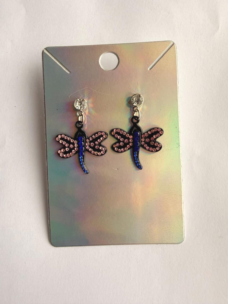 May include: A pair of dragonfly earrings featuring black metal frames, blue crystal bodies, and pink crystal wings. Each earring hangs from a clear round stud and is displayed on a rectangular, iridescent silver-toned cardboard backing card.