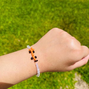 May include: A beaded bracelet featuring a central design of orange and black beads. The bracelet is made with clear, iridescent beads and has a silver-colored clasp. The background is a blurred green lawn.