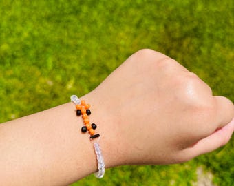 May include: A beaded bracelet featuring a central design of orange and black beads. The bracelet is made with clear, iridescent beads and has a silver-colored clasp. The background is a blurred green lawn.