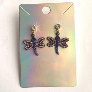 May include: A pair of dragonfly earrings featuring black metal bodies with blue accents and pink rhinestone wings. Each earring is topped with a single clear round rhinestone stud and is displayed on a rectangular iridescent card.
