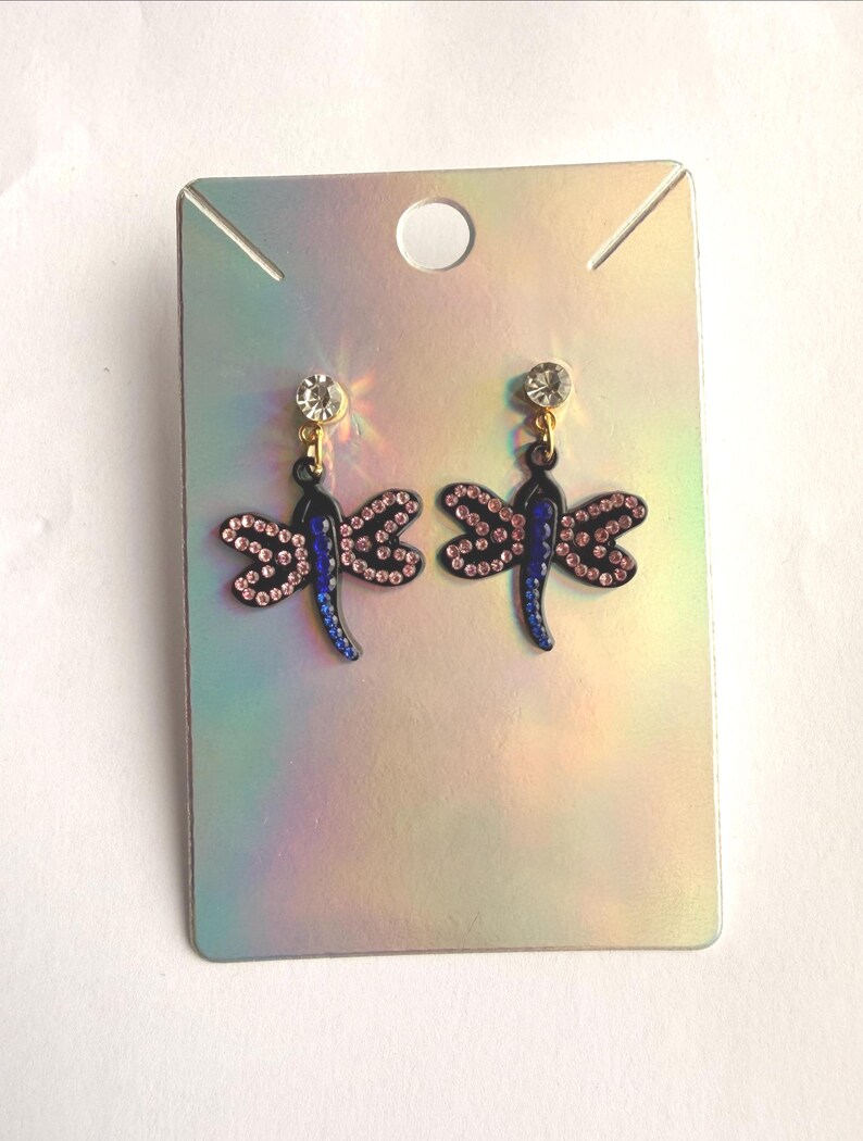 May include: A pair of dragonfly earrings featuring black metal bodies with blue accents and pink rhinestone wings. Each earring is topped with a single clear round rhinestone stud and is displayed on a rectangular iridescent card.