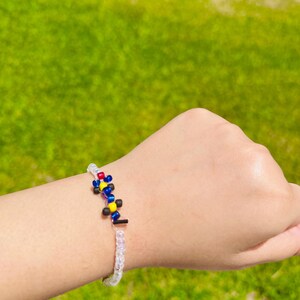 May include: A beaded bracelet featuring a central design of red, blue, yellow, and black beads. The bracelet is strung on a clear, elastic cord, and is displayed against a backdrop of green grass.