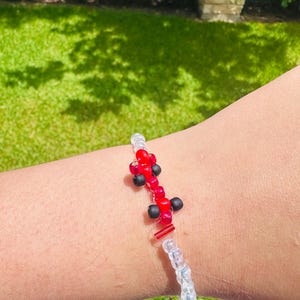 May include: A beaded bracelet featuring red and black beads in a unique design. The bracelet has a clear beaded section and a small gold-colored clasp. The bracelet is shown on a person's arm against a background of green grass.