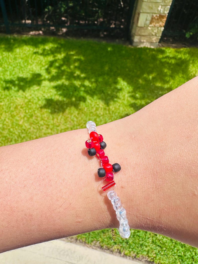 May include: A beaded bracelet featuring red and black beads in a unique design. The bracelet has a clear beaded section and a small gold-colored clasp. The bracelet is shown on a person's arm against a background of green grass.