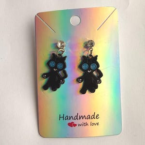 May include: A pair of dangle earrings featuring black cat charms with light blue button eyes, attached to clear crystal studs. The earrings are displayed on a rectangular holographic card with the text "Handmade with love" and two red hearts.