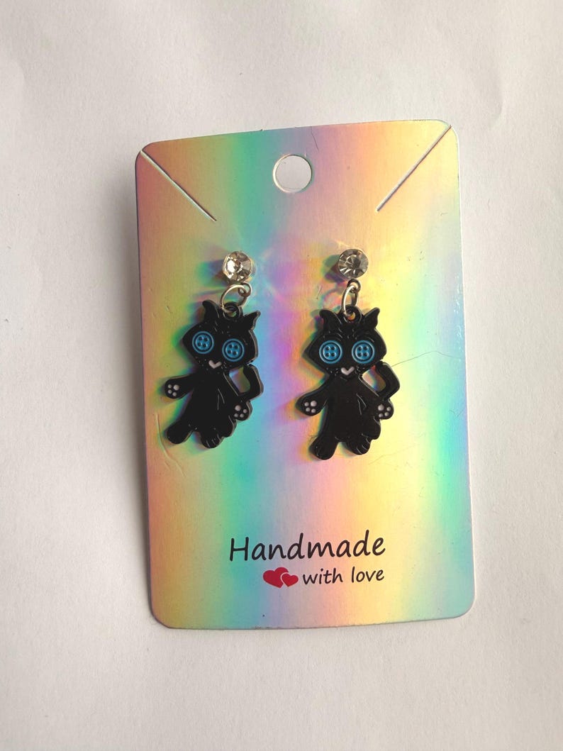 May include: A pair of dangle earrings featuring black cat charms with light blue button eyes, attached to clear crystal studs. The earrings are displayed on a rectangular holographic card with the text "Handmade with love" and two red hearts.