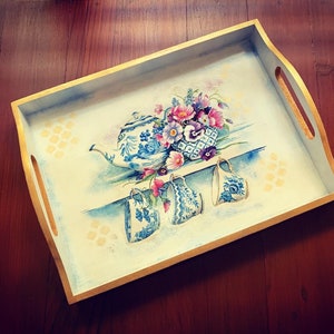 Puede incluir: Una bandeja de madera con un borde dorado, pintada con un diseño floral y una tetera con cuatro tazas. Las flores son azules, rosas y blancas. La tetera y las tazas son azules y blancas.