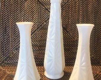 May include: Three white, vintage-style vases with geometric and leaf-like patterns. The vases vary in height and shape, with a tall, slender vase in the center and two shorter, wider vases on either side. The vases are made of a glossy white material.