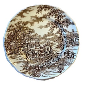 May include: A decorative plate with a brown transferware design. The plate features a detailed landscape scene with a horse-drawn carriage, buildings, and trees. The rim has a scalloped edge, and the plate is set against a white background.