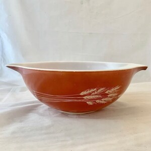 May include: A vintage, rust-colored mixing bowl with a white interior and a wheat stalk design. The bowl has a wide rim and a pouring spout. The wheat design is in white. This bowl is perfect for baking and cooking.