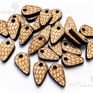 May include: A collection of teardrop-shaped wooden beads featuring a light brown wood grain surface with a repeating pattern of small, tan, oval-shaped spots. Each bead has a dark brown edge and a single circular hole drilled near the top.