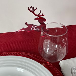 May include: Red acrylic reindeer place card with the name "Elsa" engraved on it. The reindeer is attached to a clear wine glass.