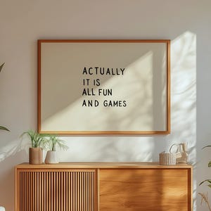Puede incluir: Un letrero enmarcado en madera con el texto "ACTUALLY IT IS ALL FUN AND GAMES" cuelga sobre un mueble de madera. Una silla y plantas están en la habitación. El mueble tiene una puerta de listones y una puerta maciza. La habitación tiene una ventana.