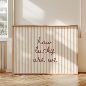 May include: A framed wall art piece with a light wood frame. The artwork features a cream and beige striped background with the words "how lucky are we" in a cursive font. The artwork is displayed in a room with windows and a wooden floor.