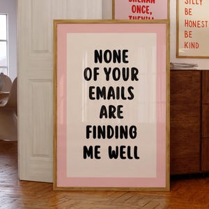 May include: A rectangular wall art print in a light wood frame featuring a pink border and a cream centre. The text in bold black sans-serif font reads: NONE OF YOUR EMAILS ARE FINDING ME WELL. The print is displayed in a room with wooden floors.