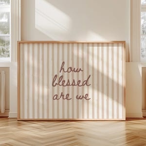May include: A framed wall art piece with a light brown wooden frame. The artwork features a striped background in beige and off-white, with the words "how blessed are we" in a cursive font.