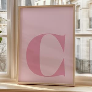 Puede incluir: Un p&oacute;ster rosa enmarcado con una gran letra rosa "C" en el centro. El p&oacute;ster est&aacute; colgado en una pared junto a una ventana con vista a una ciudad.