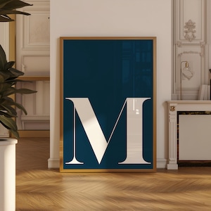 May include: A framed art print featuring a large white letter "M" against a deep navy blue background. The artwork is encased in a gold-colored frame, creating a striking contrast. The print is displayed indoors, leaning against a wall.