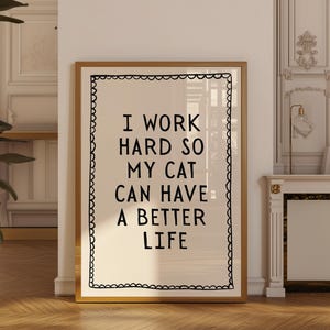 Peut inclure: Une affiche encadrée avec le texte "I WORK HARD SO MY CAT CAN HAVE A BETTER LIFE" en gras noir. L'affiche a un fond crème et une bordure noire décorative, le tout dans un cadre doré.