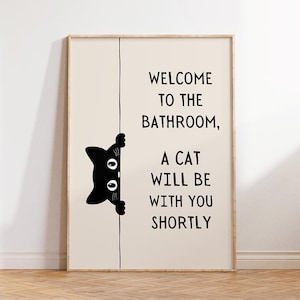 May include: A rectangular wall print with a light beige background featuring a black graphic of a cat peeking from behind a vertical line. The text reads: WELCOME TO THE BATHROOM, A CAT WILL BE WITH YOU SHORTLY in a simple black sans-serif font.