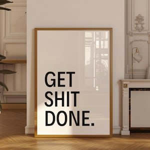 Op de afbeelding: Een ingelijste print met de tekst "GET SHIT DONE" in vette zwarte letters op een witte achtergrond. De lijst heeft een warme goudkleur en de print wordt weergegeven tegen een witte muur met een houten vloer.