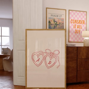 Puede incluir: Una impresión enmarcada con dos medallones rojos en forma de corazón unidos por un lazo, con las letras "A" y "D". La impresión se exhibe con otras dos obras de arte enmarcadas, una con el texto "CONSIDER IT ALL JOY" y otra con una pintura de paisaje.