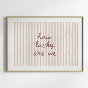 May include: Framed art print with a gold-colored frame. The artwork features a striped background in beige and white, with the phrase "how lucky are we" in a cursive, reddish-brown font. A decorative piece for home decor.