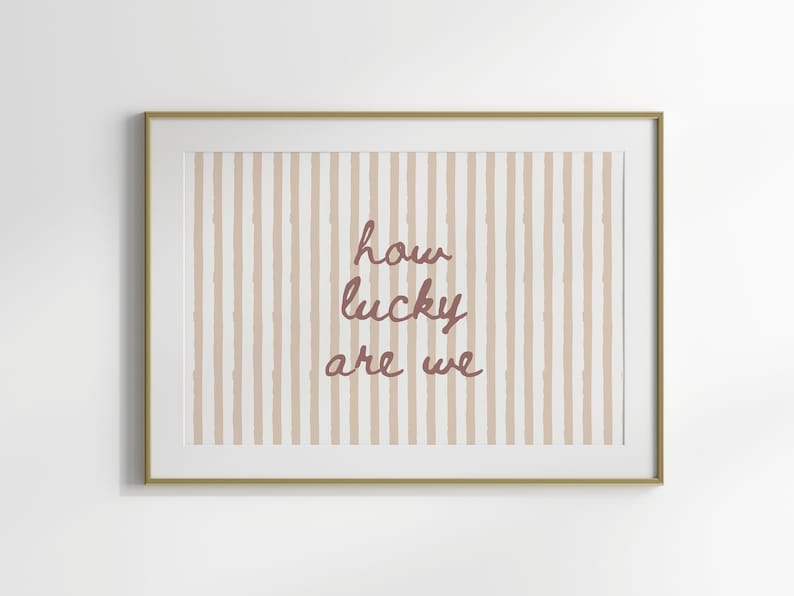 May include: Framed art print with a gold-colored frame. The artwork features a striped background in beige and white, with the phrase "how lucky are we" in a cursive, reddish-brown font. A decorative piece for home decor.