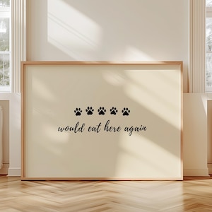 May include: A framed print with a light beige background. Four black paw prints are above the phrase "would eat here again" in a cursive font. The frame is a light brown color. The print is displayed on a wooden floor.