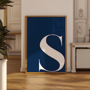 May include: A framed art print featuring a large, elegant letter "S" in a cream color against a deep navy blue background. The artwork is encased in a gold-colored frame, adding a touch of sophistication to the design. The print is displayed on a wooden floor.