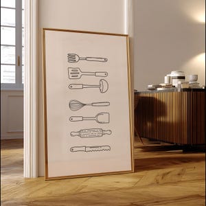 May include: A framed print featuring line drawings of various kitchen utensils, including a fork, spatula, ladle, whisk, turner, rolling pin, and knife. The artwork is in black and white, with a gold frame, and leans against a white wall.