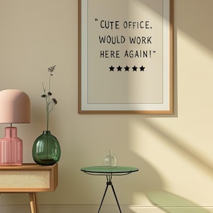 May include: A framed print with the text "CUTE OFFICE, WOULD WORK HERE AGAIN!" and five stars. The image also features a pink lamp, a green vase, a small side table, and a patterned rug.