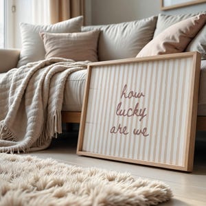 May include: A framed wall art piece with the text "how lucky are we" in a cursive font. The artwork has a striped pattern in beige and white, and is leaning against a beige sofa. A fluffy rug and throw blanket are also visible.