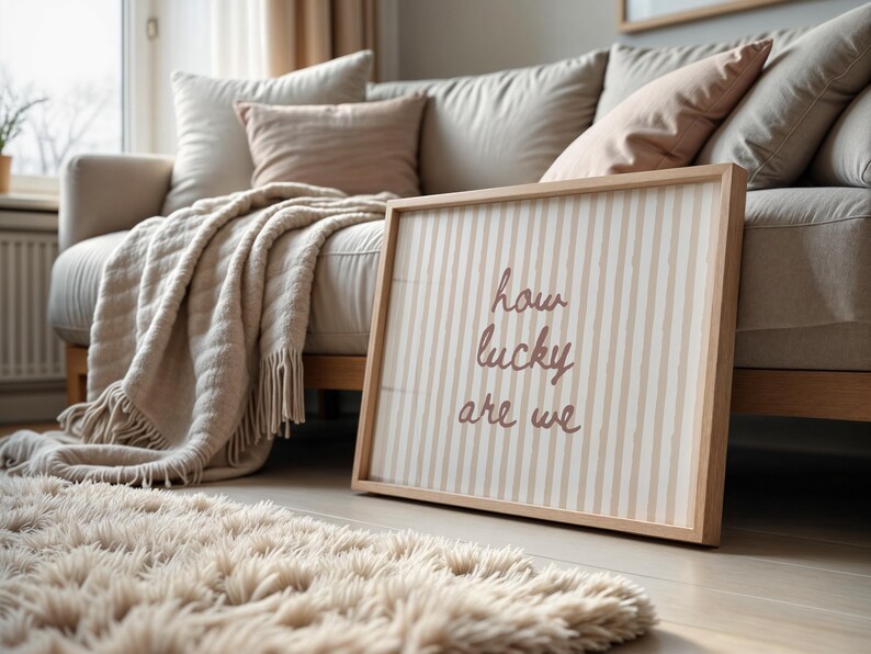 May include: A framed wall art piece with the text "how lucky are we" in a cursive font. The artwork has a striped pattern in beige and white, and is leaning against a beige sofa. A fluffy rug and throw blanket are also visible.