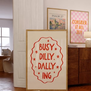 May include: Three framed art prints. The largest print has the words "BUSY DILLY DALLY ING" in red on a white background. Other prints feature a landscape and a pink and white checkerboard with the words "CONSIDER IT ALL JOY".