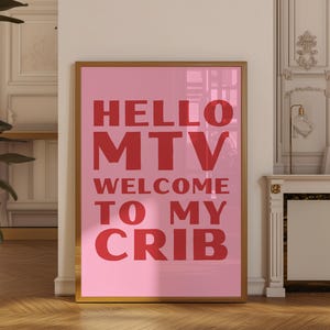 Puede incluir: Un póster rosa enmarcado con las palabras "HELLO MTV WELCOME TO MY CRIB" en letras rojas. El póster está en un marco dorado y se muestra contra una pared blanca.