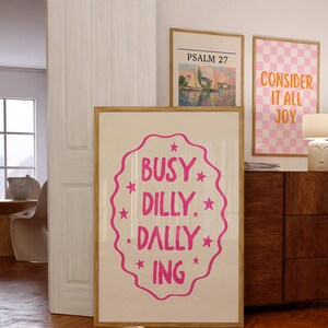 May include: Three framed art prints. The largest print has the words "BUSY DILLY DALLY ING" in pink. The other prints feature a landscape and the words "CONSIDER IT ALL JOY". All prints have wooden frames.