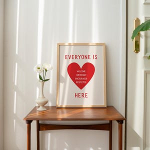 May include: A framed print with the words "EVERYONE IS HERE" and a red heart graphic. The heart contains the words "WELCOME, IMPORTANT, ENCOURAGED, RESPECTED". The print is on a wooden table with a vase of white flowers.