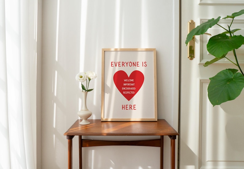 May include: A framed print with the words "EVERYONE IS HERE" and a red heart graphic. The heart contains the words "WELCOME, IMPORTANT, ENCOURAGED, RESPECTED". The print is on a wooden table with a vase of white flowers.