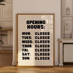 以下が含まれることがあります： 「OPENING HOURS:」のテキストが記載されたフレーム付きプリント。各曜日の後に「CLOSED」と記載されています。フレームは金色で、背景はオフホワイトです。