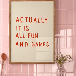May include: A framed print with the words "ACTUALLY IT IS ALL FUN AND GAMES" in red lettering. The print is in a light wood frame and hangs against a pink tiled wall. A wooden shelf below holds a glass jar and a vase.