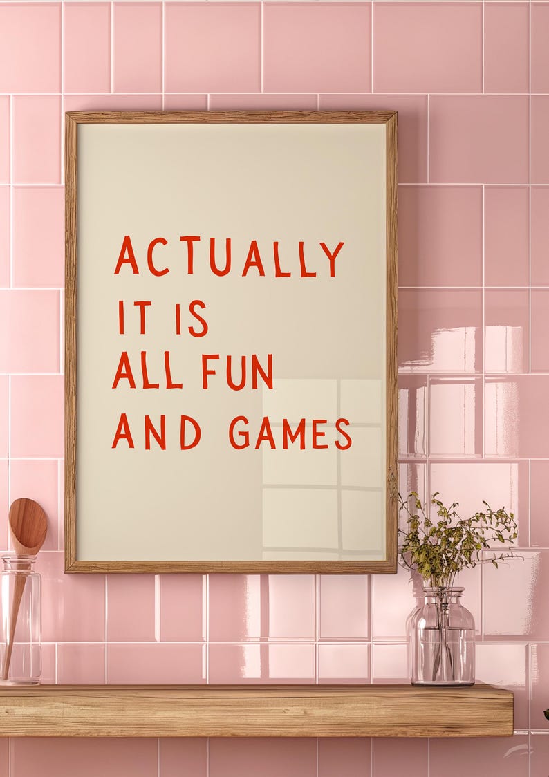 May include: A framed print with the words "ACTUALLY IT IS ALL FUN AND GAMES" in red lettering. The print is in a light wood frame and hangs against a pink tiled wall. A wooden shelf below holds a glass jar and a vase.