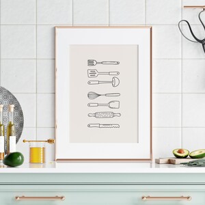 May include: A framed print featuring line drawings of kitchen utensils, including a fork, spatula, ladle, whisk, spoon, rolling pin, and knife. The artwork is displayed on a white tiled wall above a mint green cabinet with copper hardware. Additional kitchen items include a plate, olive oil bottles, and avocados.