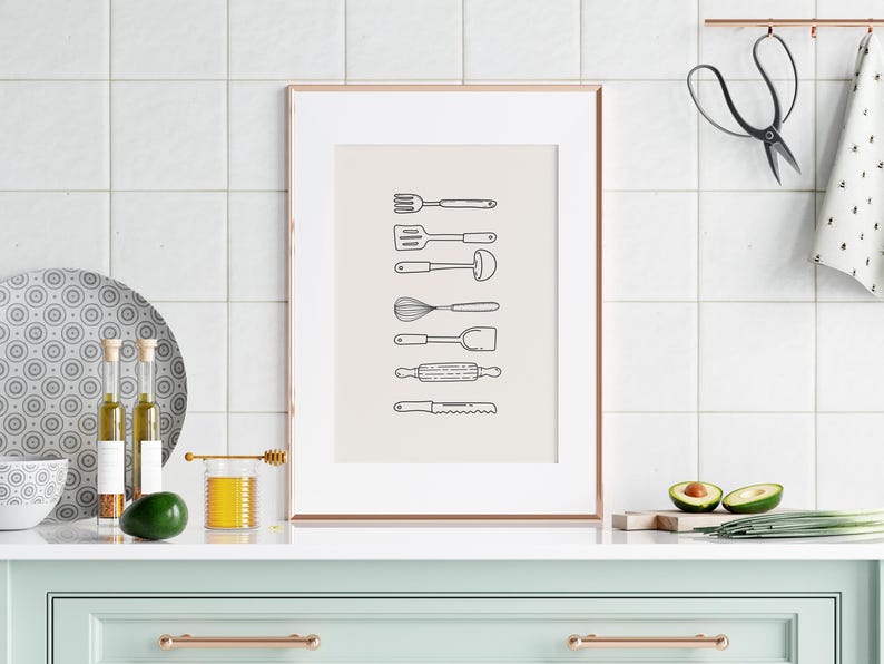 May include: A framed print featuring line drawings of kitchen utensils, including a fork, spatula, ladle, whisk, spoon, rolling pin, and knife. The artwork is displayed on a white tiled wall above a mint green cabinet with copper hardware. Additional kitchen items include a plate, olive oil bottles, and avocados.