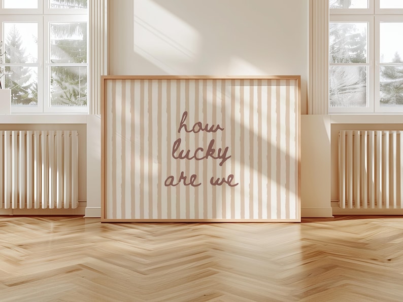 May include: A framed wall art piece with a light wood frame. The artwork features a cream and beige striped background with the words "how lucky are we" in a cursive font. The artwork is displayed in a room with windows and a wooden floor.