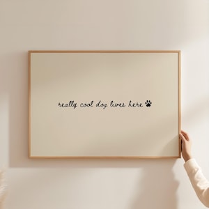 以下が含まれることがあります： クリーム色の背景と木製フレームの額入りプリント。 「really cool dog lives here」というテキストが筆記体で書かれ、黒い足跡が続きます。 人がプリントを持っています。