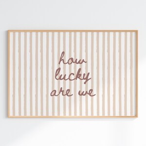 May include: A framed print with a light brown wooden frame. The artwork features a striped background in beige and white, with the words "how lucky are we" in a cursive, reddish-brown font.