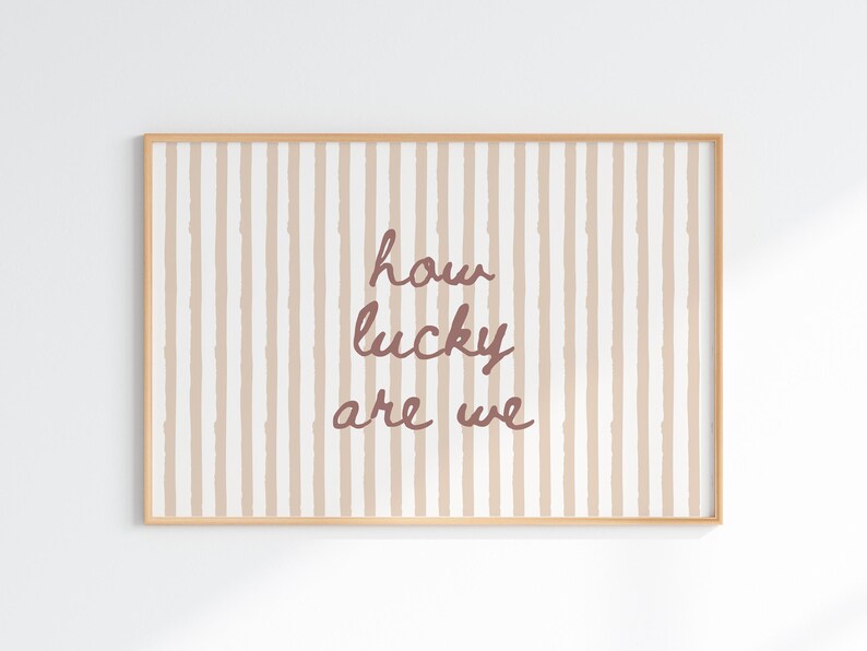 May include: A framed print with a light brown wooden frame. The artwork features a striped background in beige and white, with the words "how lucky are we" in a cursive, reddish-brown font.