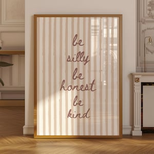 May include: A rectangular wall art print in a gold frame featuring vertical beige and white stripes. The text "be silly be honest be kind" is printed in a dark brown, handwritten-style font centered on the striped background.