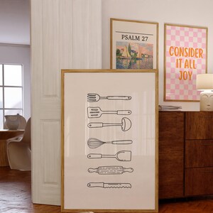 May include: A framed print featuring line drawings of kitchen utensils, including a fork, spatula, ladle, whisk, spoon, rolling pin, and knife. The print is in a wooden frame and is displayed with other framed art.